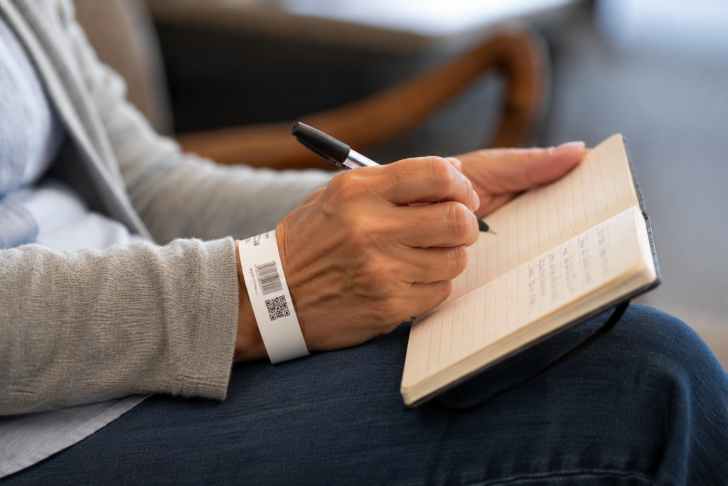Close-up of a patient's hand writing in a notebook, a hospital ID wristband visible on their wrist.